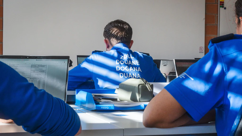 Plusieurs agents des douanes en uniforme bleu sont assis devant des ordinateurs dans une salle de formation. Sur le dos d’un agent figurent les mots Zoll, Douane, Dogana et Duana. La scène montre une situation d’apprentissage en intérieur.  
