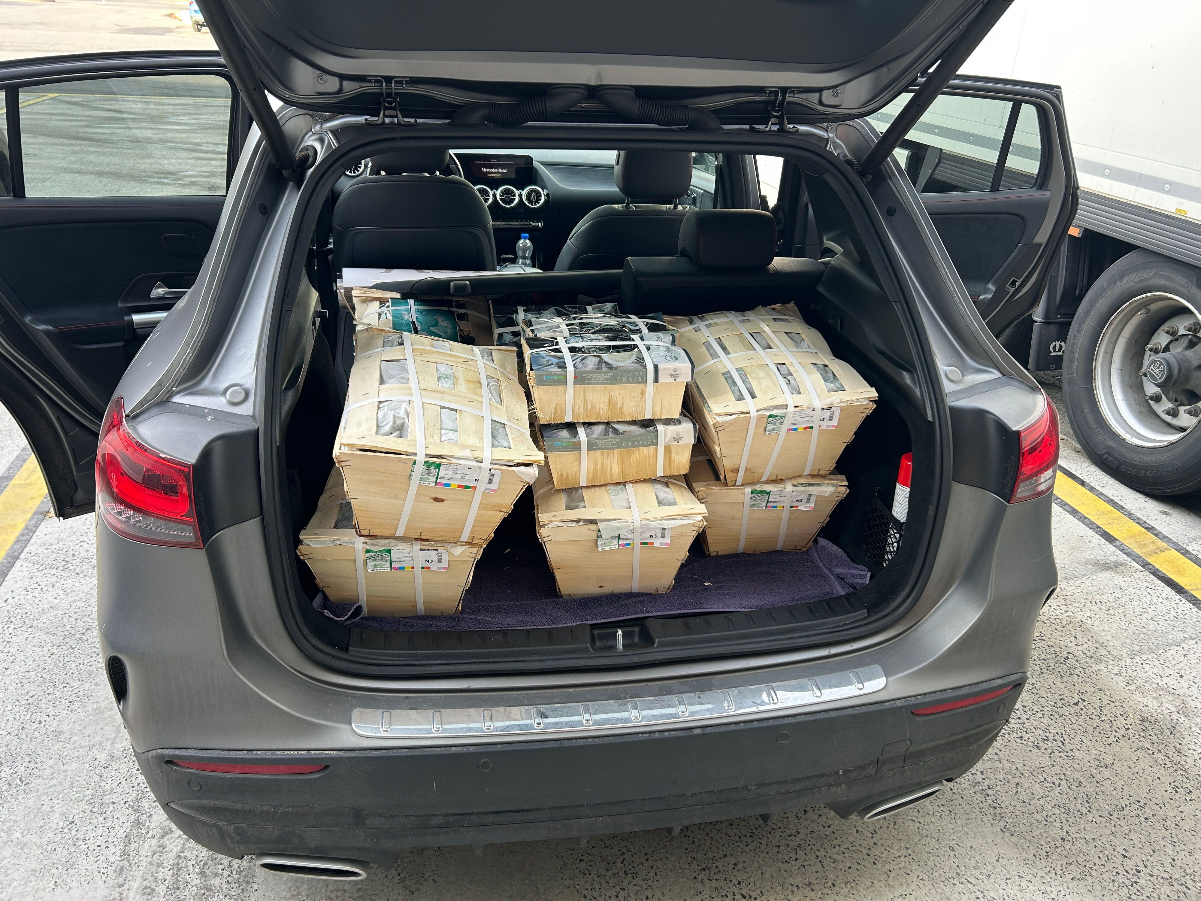 Une voiture avec le coffre ouvert, remplie de plusieurs caisses contenant apparemment des huîtres. Les caisses sont sécurisées avec du ruban adhésif et empilées dans le véhicule.