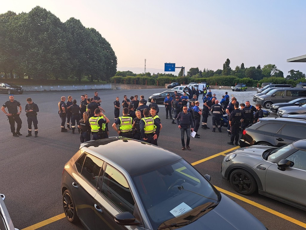 Un groupe d'agents des douanes se rassemble sur un parking à la frontière. Plusieurs véhicules sont visibles en arrière-plan. La scène se déroule en plein jour.