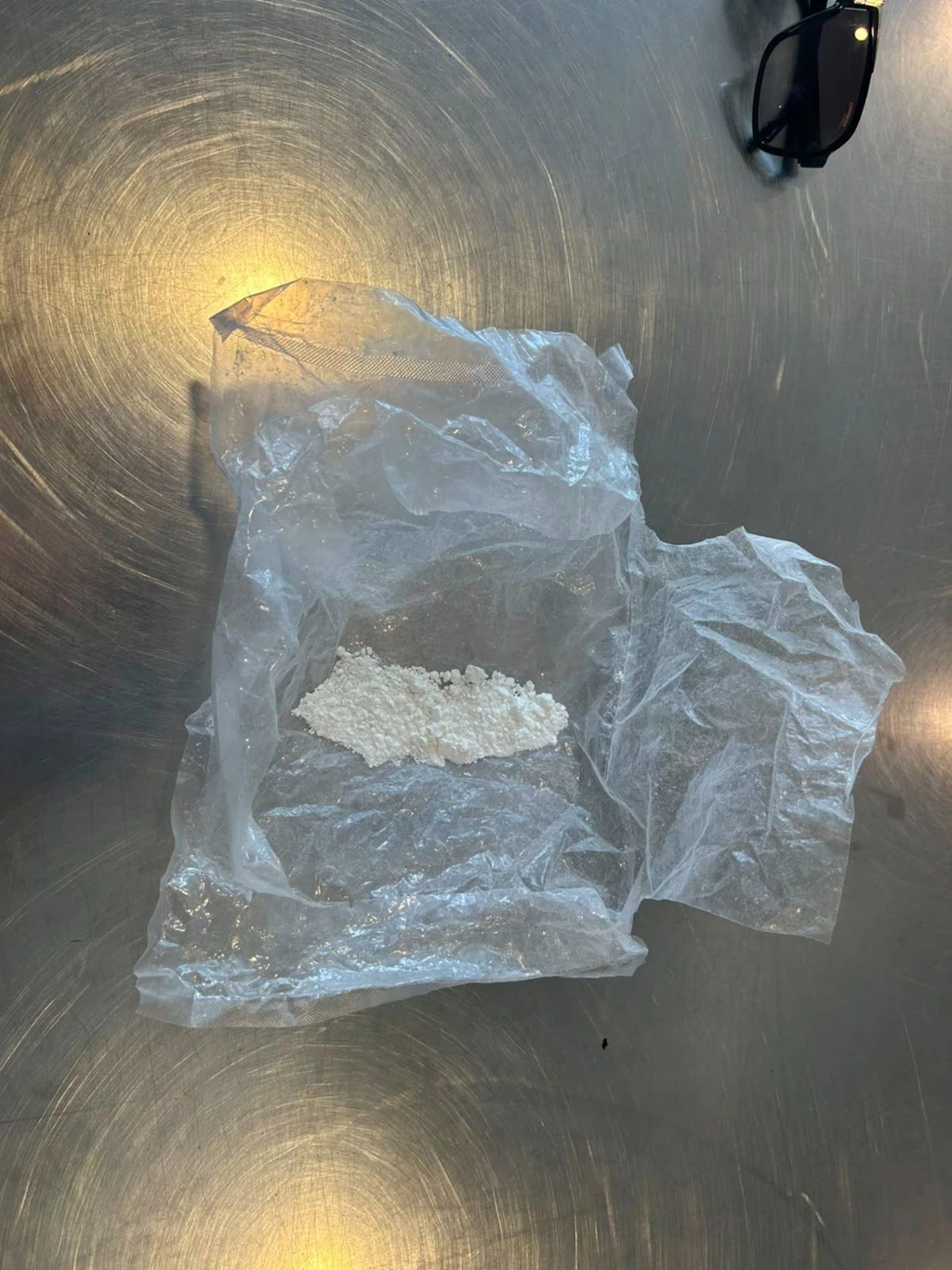 Sur une table métallique se trouve un sachet en plastique transparent contenant une petite quantité de poudre blanche, probablement de la cocaïne. Dans le coin supérieur droit, on aperçoit des lunettes de soleil. L’image illustre une scène typique de contrôle antidrogue.