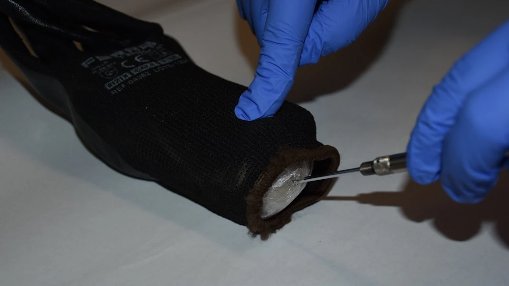 Une paire de mains gantées examine un gant noir contenant un paquet emballé dans du film plastique. Un outil est utilisé pour dévoiler le paquet, probablement lors d'une saisie de drogue. La scène se déroule sur une surface claire.  