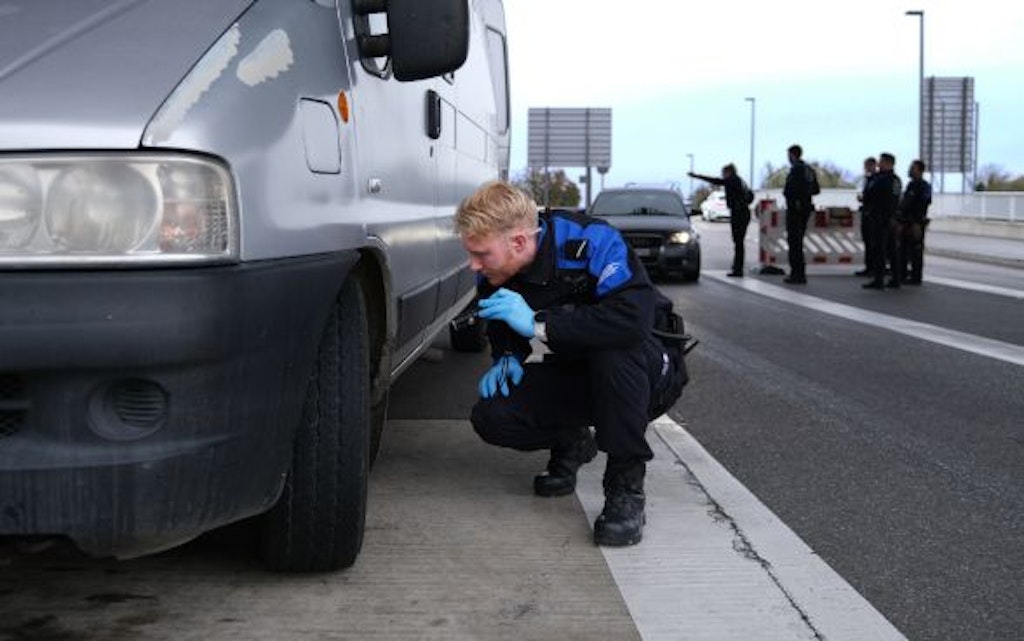 Un agent des douanes inspecte un véhicule sur une route. En arrière-plan, d'autres agents et véhicules sont visibles. La scène suggère un contrôle routier.