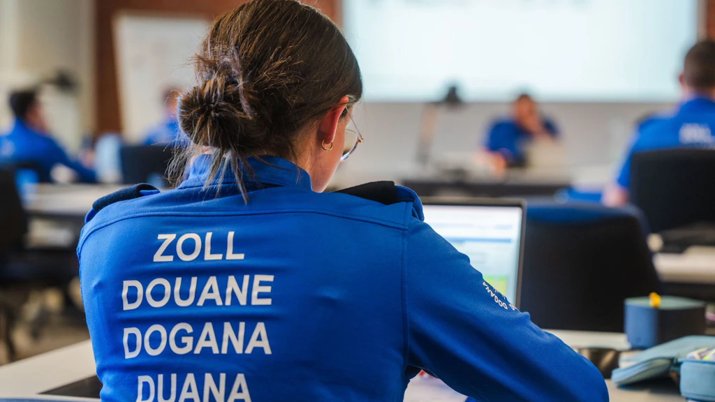  Une personne en uniforme bleu portant l'inscription "Zoll, Douane, Dogana, Duana" est assise à une table et travaille sur un ordinateur portable. À l'arrière-plan, d'autres personnes en uniforme similaire participent à une formation.