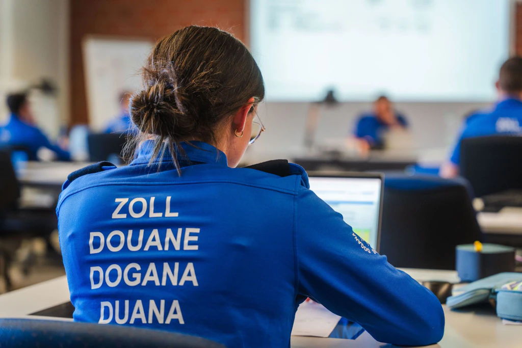  Une personne en uniforme bleu portant l'inscription "Zoll, Douane, Dogana, Duana" est assise à une table et travaille sur un ordinateur portable. À l'arrière-plan, d'autres personnes en uniforme similaire participent à une formation.