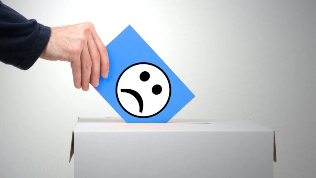Sur cette image, quelqu'un jette une carte bleue avec un smiley triste dans un carton gris. Ce carton est fermé et possède une fente comme une boîte aux lettres. Le carton est placé devant un mur gris. On ne voit de la personne que la main droite et une partie du bras, recouverte d'une manche bleu foncé.