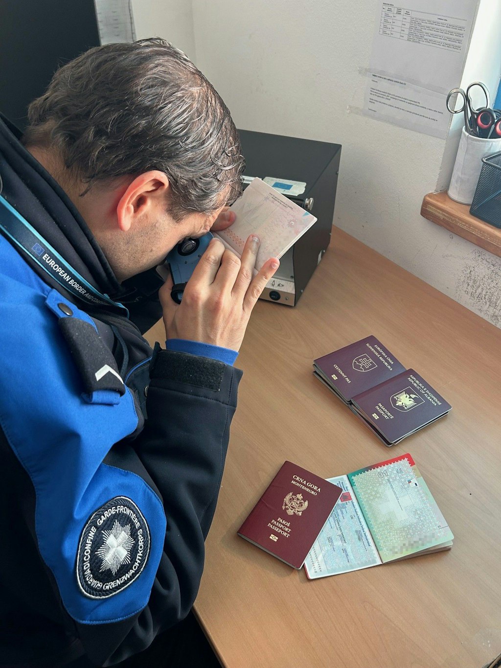 Un agent des douanes et des frontieres verifie l authenticite d un passeport a l aide d un appareil sur un bureau. Devant lui se trouvent plusieurs passeports et documents.  