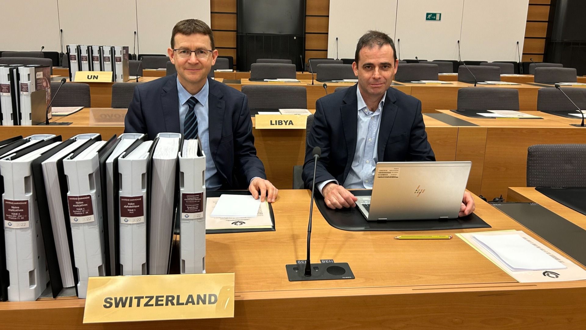 Deux hommes sont assis à une table de conférence avec l'inscription "Switzerland". Devant eux se trouvent plusieurs classeurs.  