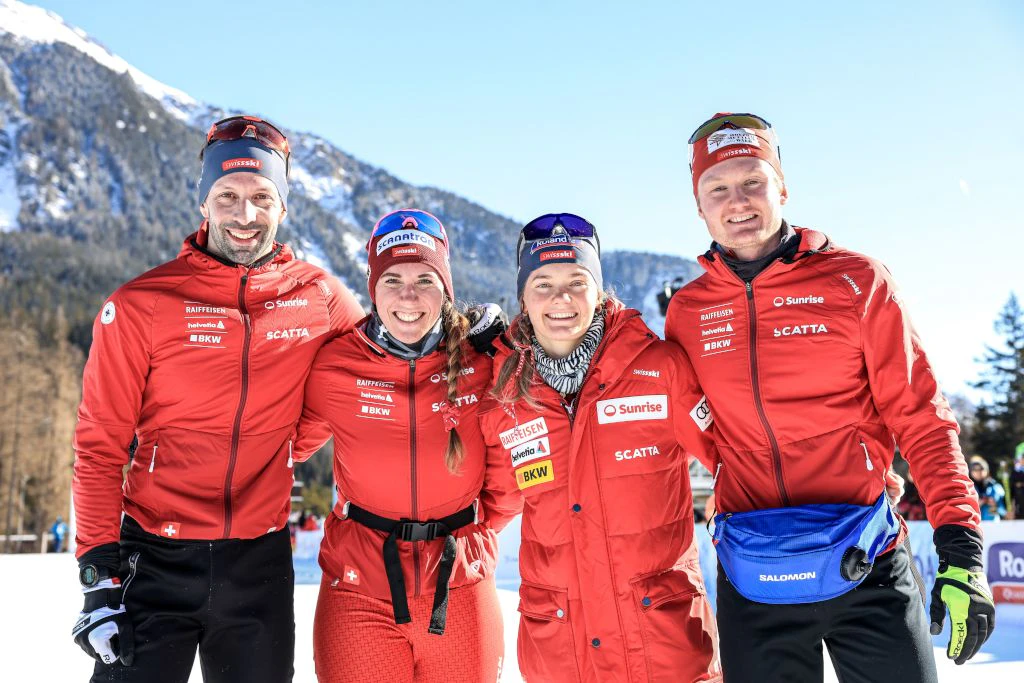 Quatre membres de l’équipe suisse de biathlon posent côte à côte dans la neige, tous portent des vestes rouges et sourient à l’appareil photo. À l’arrière-plan, on voit des montagnes enneigées et un ciel bleu.