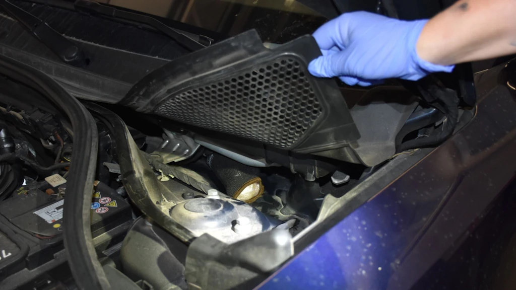 Une personne portant des gants bleus soulève un couvercle dans le compartiment moteur d'une voiture pour inspecter une zone en dessous. Divers composants du moteur et une partie de la batterie sont visibles. La scène suggère un contrôle ou une maintenance.