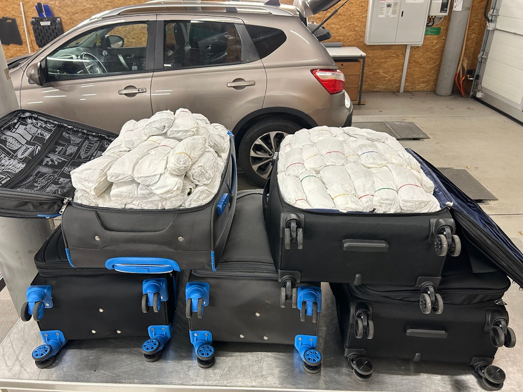 Plusieurs valises remplies de paquets de Khat sont ouvertes dans un garage. Une voiture est visible en arrière-plan. 