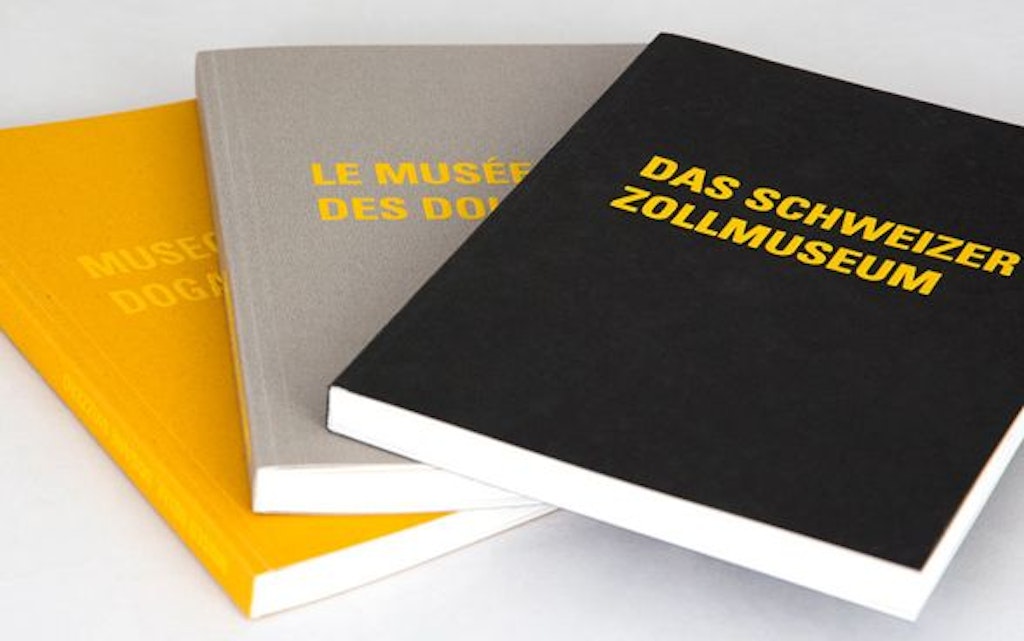 Trois livres avec les titres "Das Schweizer Zollmuseum", "Le Musée des Douanes" et "Museo delle Dogane" sont posés sur une table. Les livres ont des couleurs différentes : noir, gris et jaune.