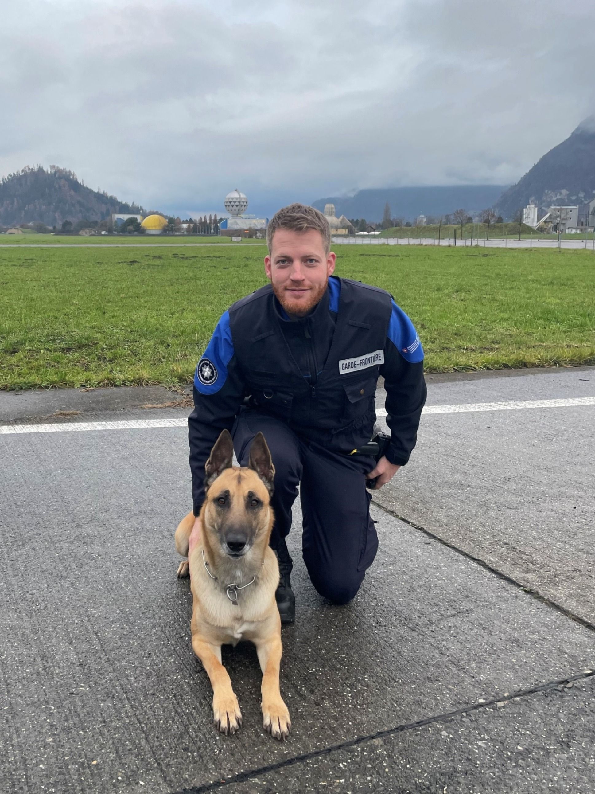 Una guardia di confine inginocchiata accanto al suo cane Malinois su una strada. Sullo sfondo, un prato verde e un cielo nuvoloso.  