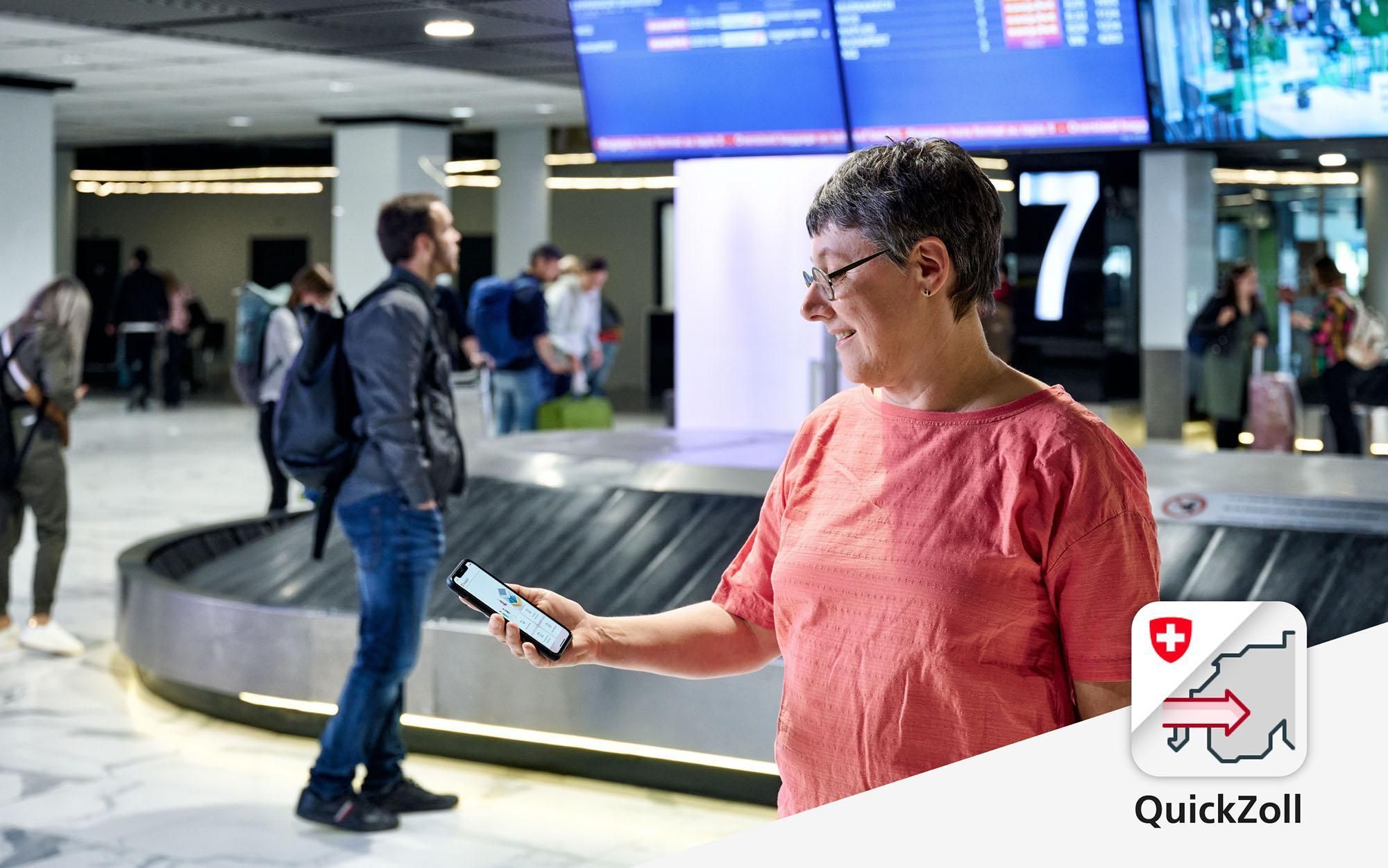Una donna nell'area bagagli di un aeroporto tiene uno smartphone con l'app QuickZoll. Intorno a lei ci sono altri viaggiatori e un grande tabellone informativo.