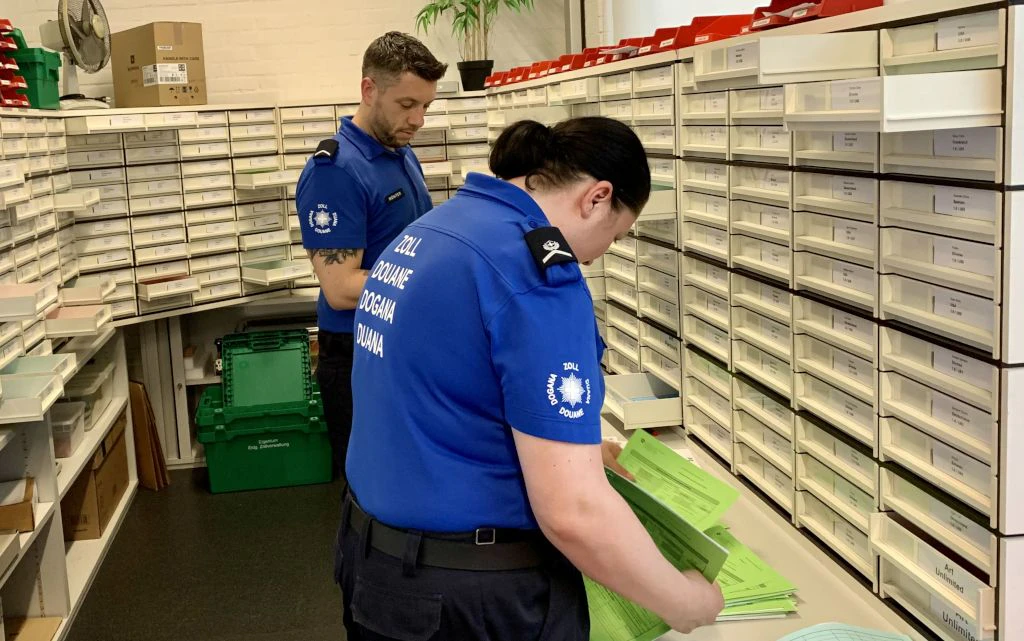 Due impiegati della dogana in uniforme blu ordinano moduli verdi in un ufficio pieno di cassetti etichettati. Sullo sfondo si vedono scaffali con documenti e materiale.  