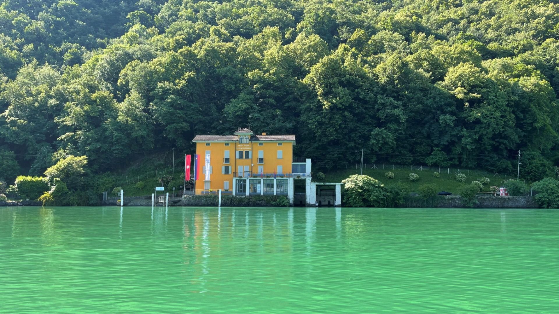 Un edificio giallo sulla riva di un lago, circondato da foresta, con bandiere svizzere.