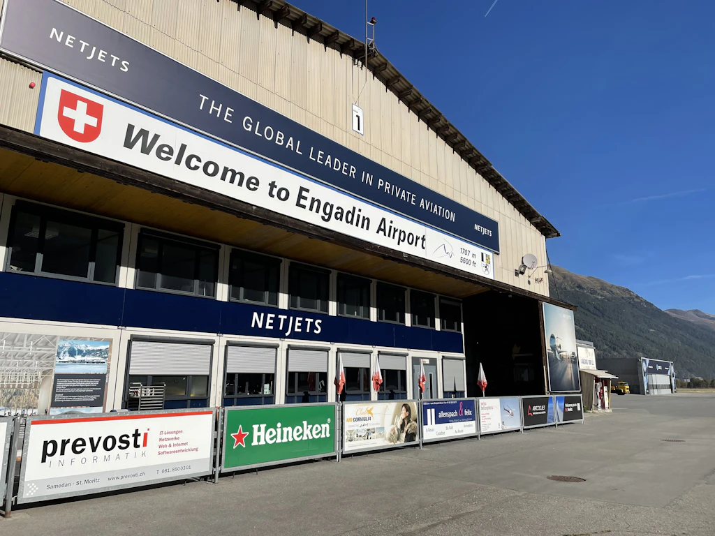Vista del terminal dell Engadin Airport con una grande insegna di ben-venuto e la croce svizzera sulla facciata. Sullo sfondo si vedono han-gar e il paesaggio montano dell Engadina.  
