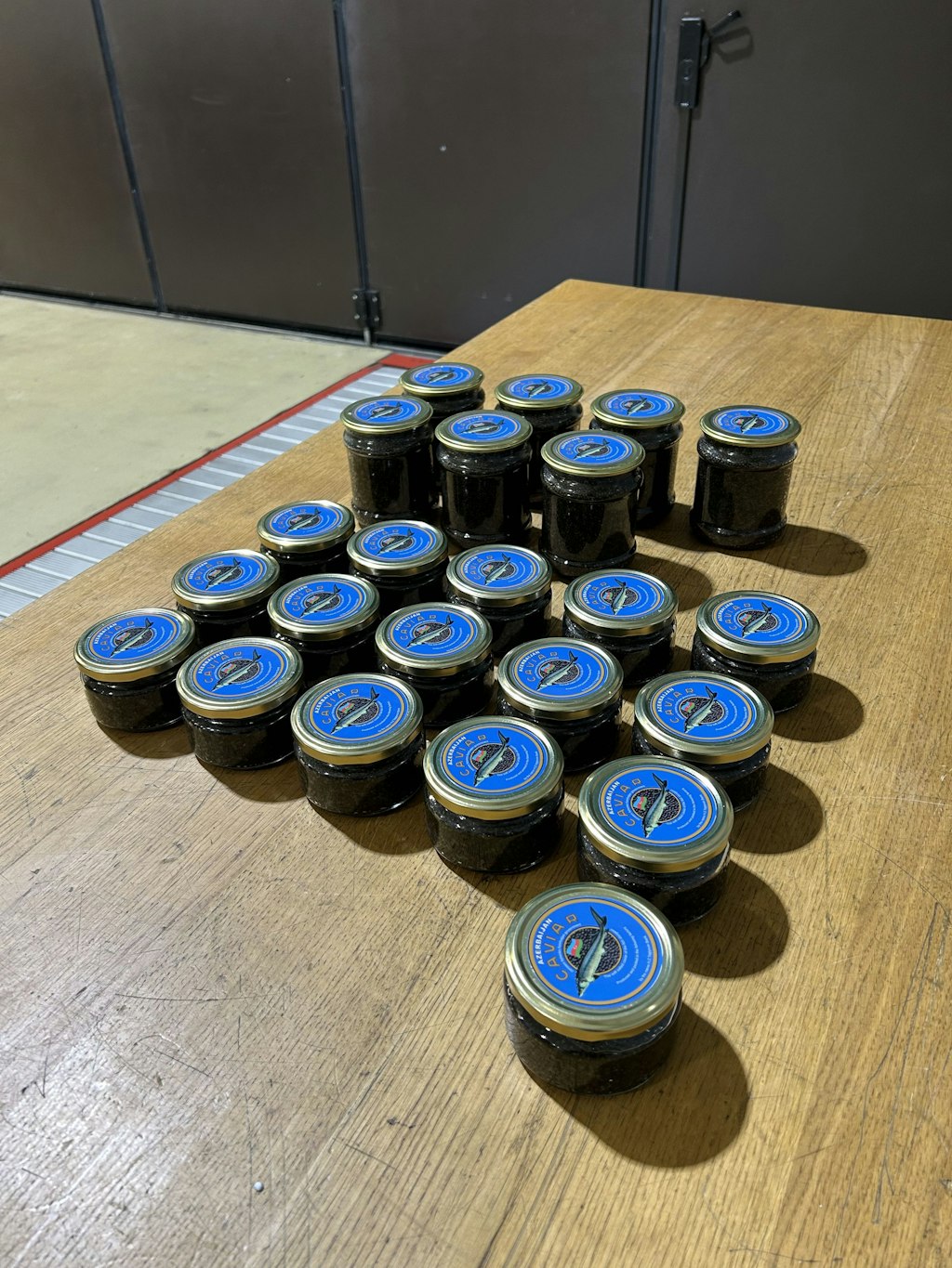 Diversi piccoli vasetti di caviale con coperchio blu sono raggruppati su un tavolo di legno in un magazzino. I vasetti sono disposti ordinatamente e sullo sfondo si vede un grande portone scuro.