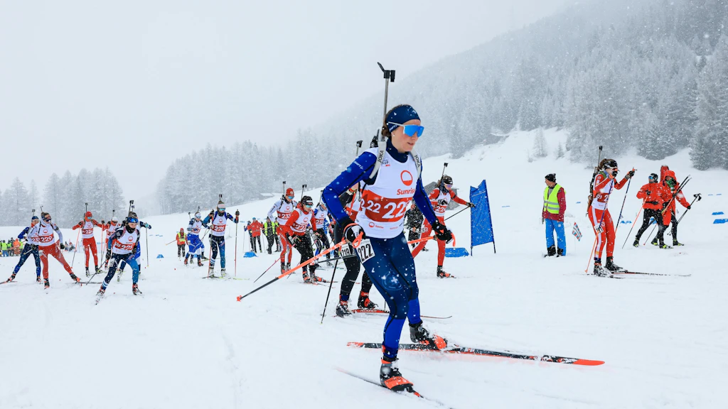Diverse biatlete e biatleti partono per una gara sotto una fitta nevicata a Ulrichen. In primo piano una atleta in tuta blu con il fucile sulla schiena scia davanti ad altri concorrenti, assistenti e un bosco innevato. 