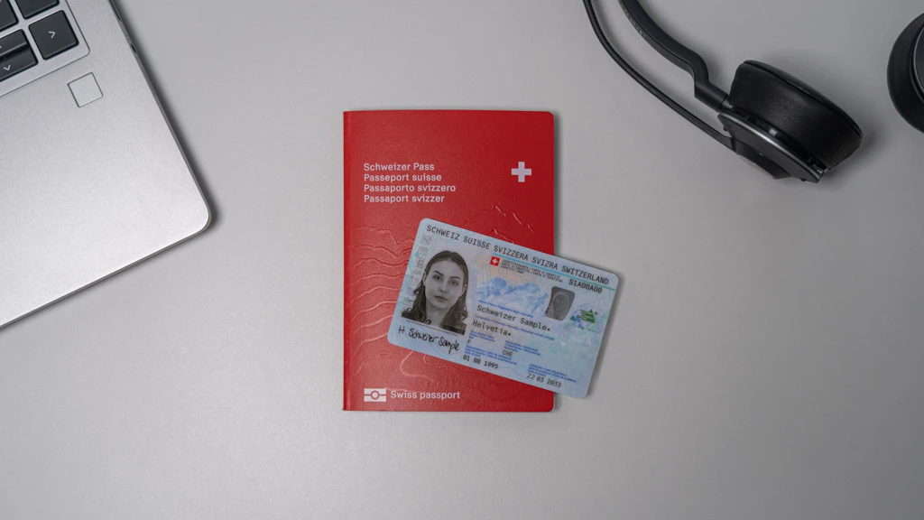 Su un tavolo chiaro ci sono un passaporto svizzero rosso e una carta d'identità svizzera. I documenti sono parzialmente circondati da un laptop e da una cuffia nera. La carta d'identità è posata sul passaporto e mostra un'immagine di esempio.  