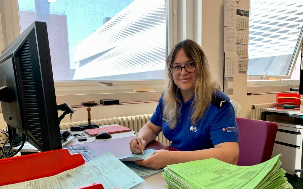 Una collaboratrice della dogana in uniforme blu e seduta a una scrivania e compila dei formulari. Davanti a lei ci sono diversi dossier verdi, uno schermo del computer e materiale dufficio.  