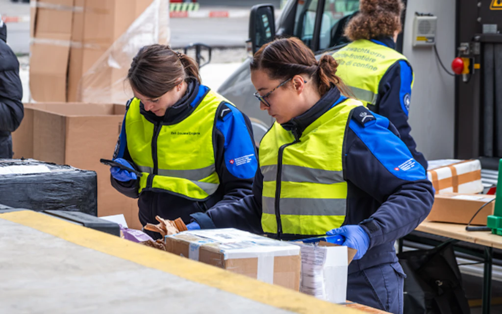 Due collaboratrici dell’Ufficio federale della dogana e della sicurezza dei confini (UDSC) controllano pacchi presso un posto doganale. Indossano uniformi e giubbotti gialli e lavorano concentrate a un tavolo con varie spedizioni. Sullo sfondo si vedono altri pacchi e un veicolo di consegna.
