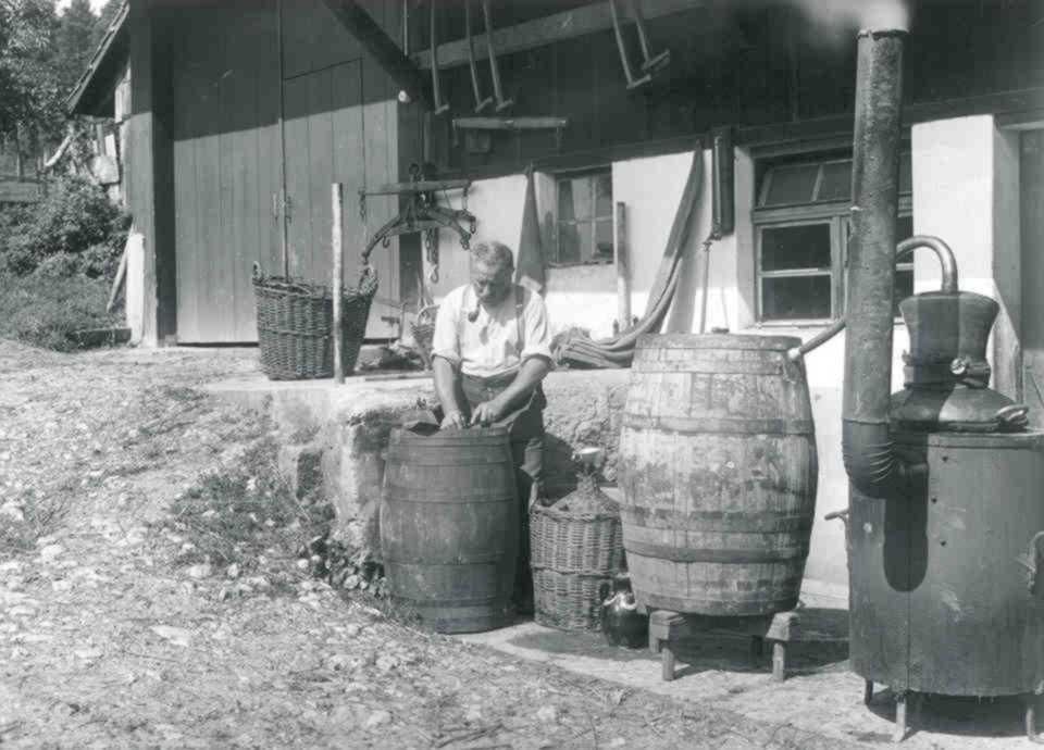 Un uomo lavora in una distilleria tradizionale all'aperto. È circondato da botti e un apparecchio di distillazione.  