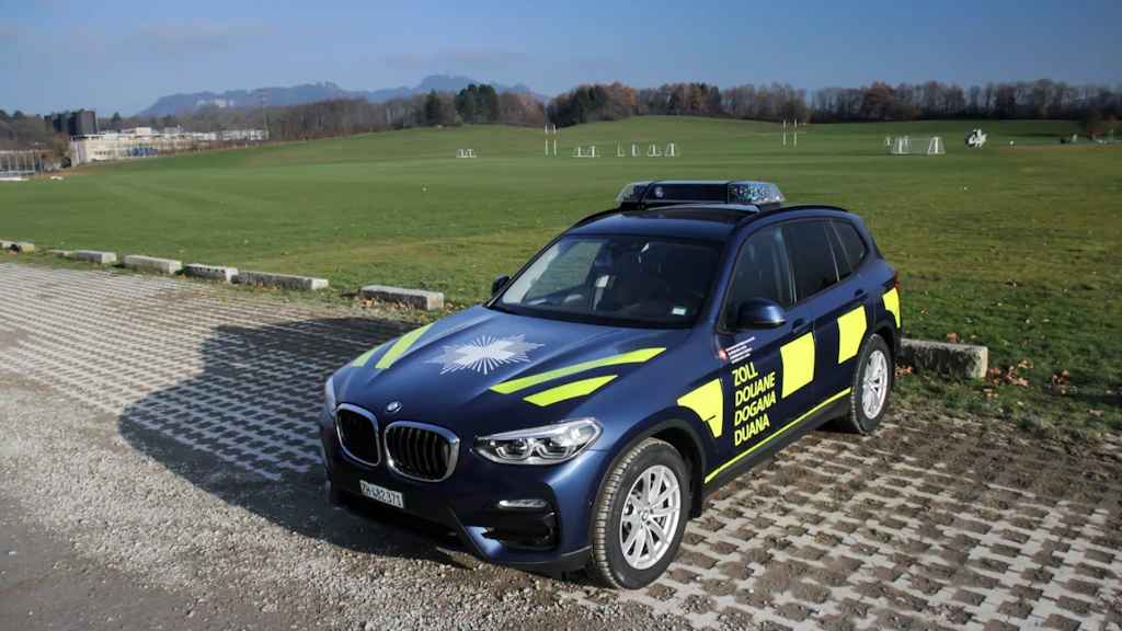 Un veicolo doganale è parcheggiato in un parcheggio. È un SUV scuro con marcature gialle e la scritta "Dogana" in diverse lingue.  