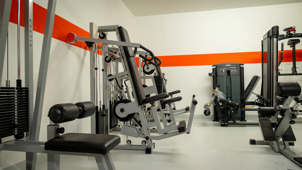 Una moderna sala fitness con diverse macchine per l'allenamento e la forza. Gli attrezzi sono disposti in una stanza luminosa con una striscia arancione sul muro. Non ci sono persone presenti.