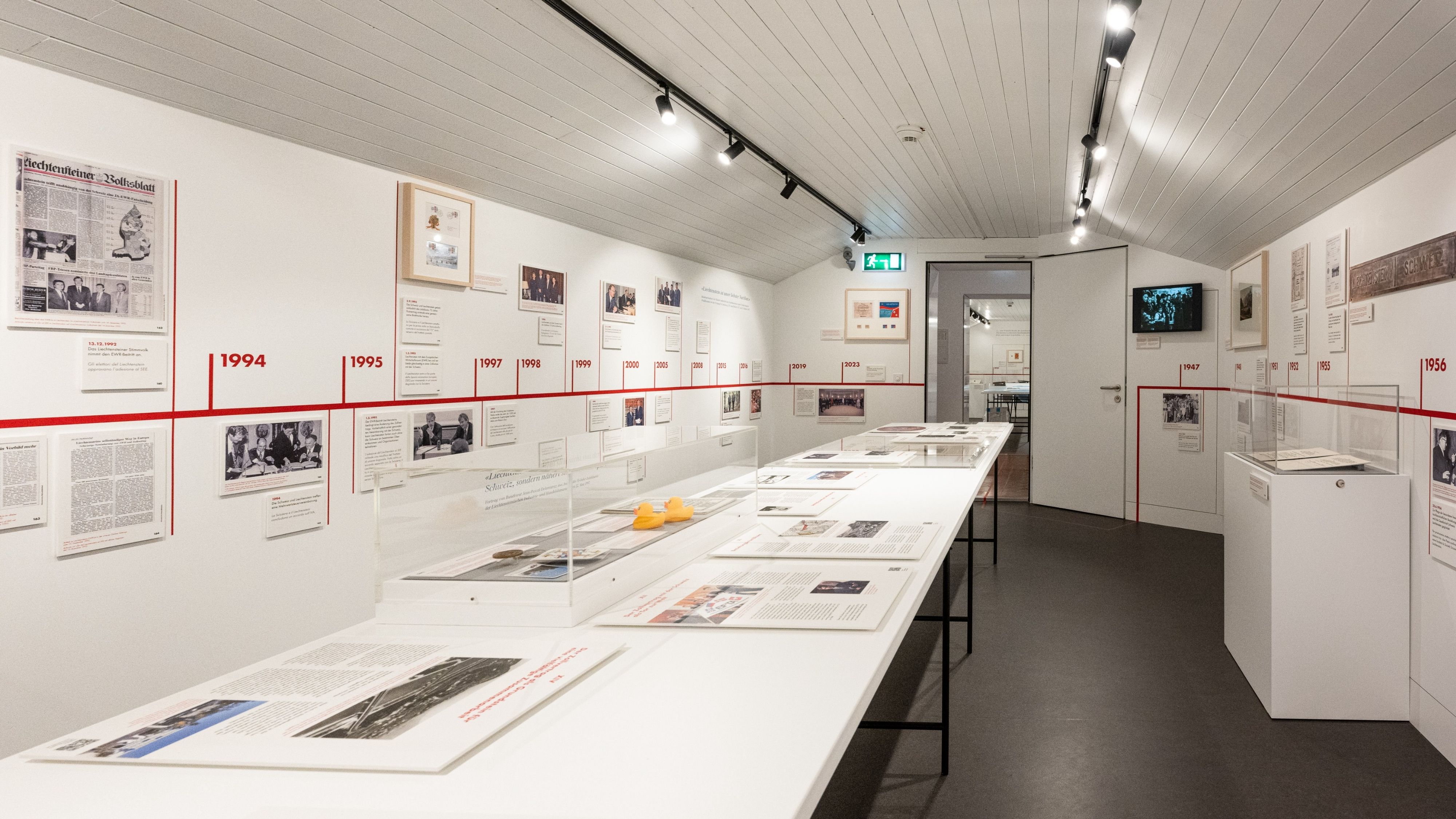 Vista di una luminosa sala espositiva del museo doganale con una lunga vetrina centrale e una linea del tempo rossa sulla parete. Documenti storici, fotografie e oggetti illustrano la storia delle dogane.