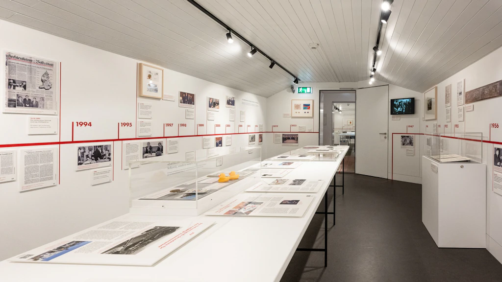 Vista di una luminosa sala espositiva del museo doganale con una lunga vetrina centrale e una linea del tempo rossa sulla parete. Documenti storici, fotografie e oggetti illustrano la storia delle dogane.