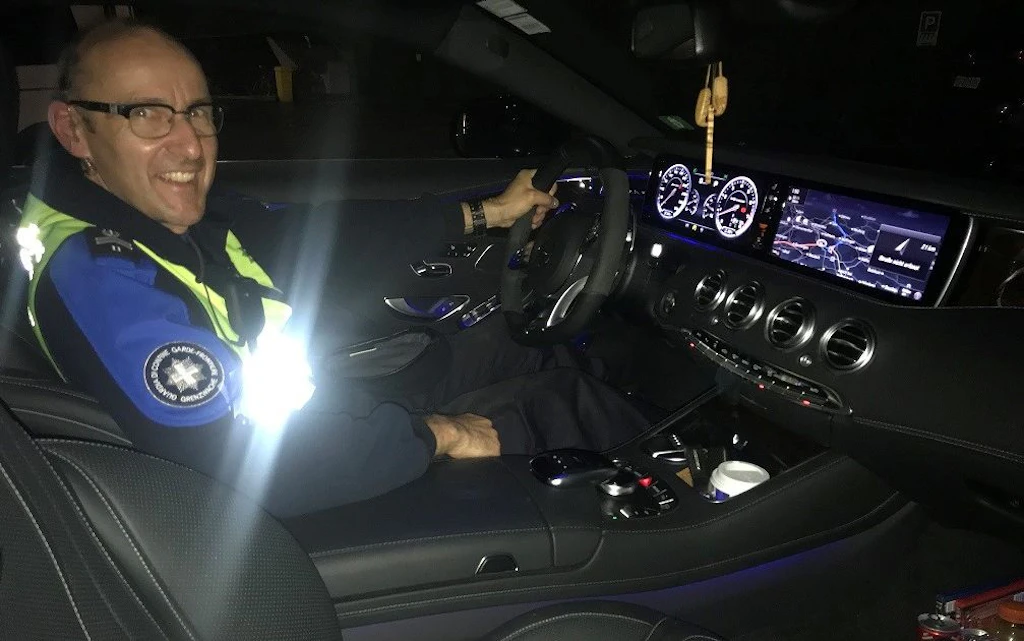 Un agente in uniforme del servizio doganale e di sicurezza delle frontiere e seduto al volante di un moderno veicolo di pattuglia di notte, con il cruscotto illuminato. Sorride verso la fotocamera mentre sullo sfondo brillano gli schermi di navigazione e del tachimetro.