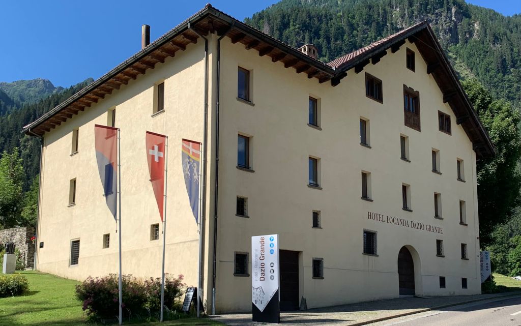 Un grande edificio tradizionale con bandiere svizzere e un cartello con la scritta "Hotel Locanda Dazio Grande" davanti a una montagna boscosa.  