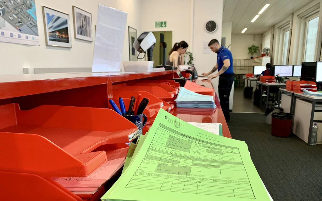 In un ufficio doganale, moduli verdi e blu sono disposti su un bancone rosso, mentre sullo sfondo il personale lavora a scrivanie e computer. La scena mostra una tipica situazione di gestione amministrativa in ambito doganale.  