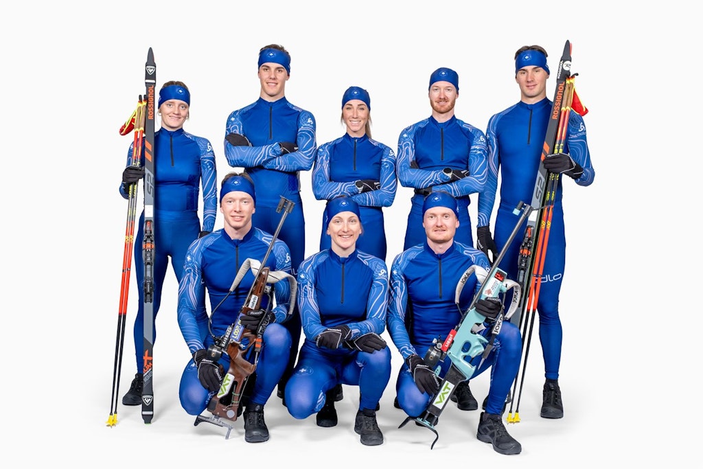 Un gruppo di nove persone in tute sportive blu posa con attrezzatura da biathlon. Portano sci e fucili e sorridono alla fotocamera.