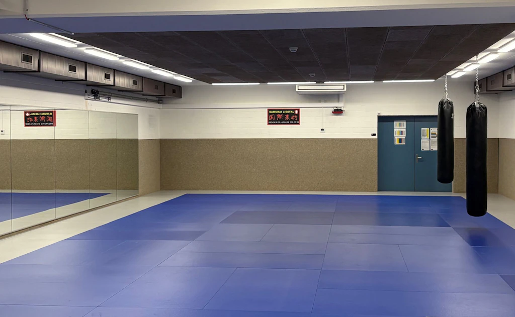 Una sala di allenamento vuota con diversi sacchi da boxe appesi e un pavimento a piastrelle rosse e blu. Sullo sfondo si vedono specchi e attrezzature da allenamento. L’illuminazione è intensa e la stanza appare pulita e ordinata.