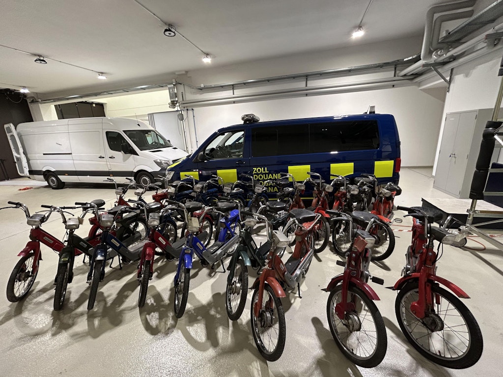 Diversi motocicli sequestrati sono disposti in un garage davanti a un veicolo doganale. Accanto c’e un furgone bianco.