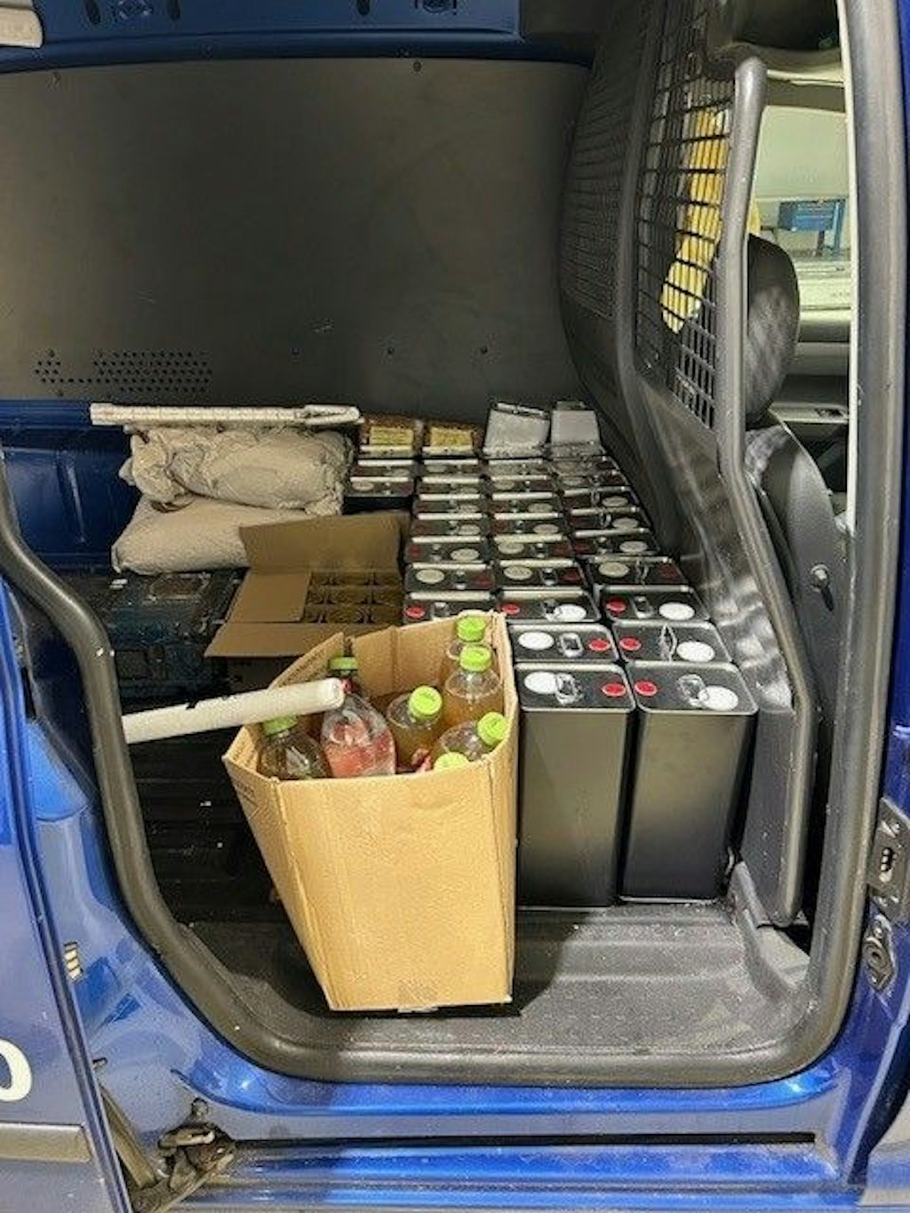 Nel vano di carico di un furgone blu si vedono numerose taniche metalliche e bottiglie di bevande in scatole di cartone, che rappresentano merce apparentemente non sdoganata. I contenitori sono disposti molto vicini tra loro e indicano una quantita considerevole di prodotti liquidi.  