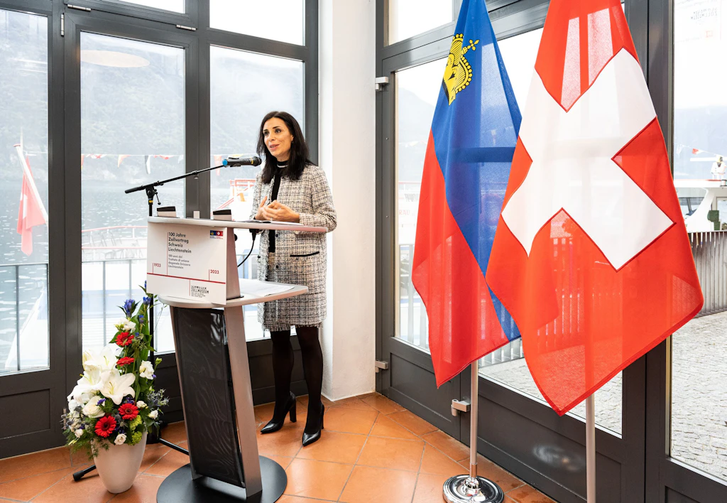 La ministra degli esteri del Principato del Liechtenstein tiene un discorso a un leggio con microfono in una sala con vista sul lago. Accanto a lei si trovano le bandiere del Liechtenstein e della Svizzera e un riferimento ai 100 anni del trattato doganale Svizzera–Liechtenstein.
