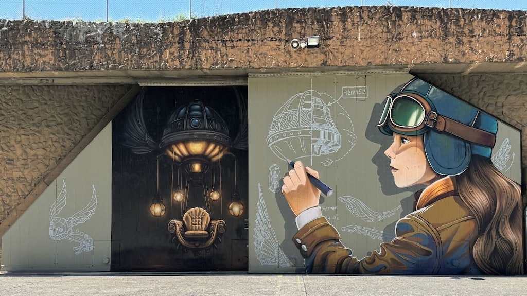 Un murale mostra una pilota con occhiali da aviatore che disegna un'astronave futuristica. Accanto, si vede una poltrona illuminata in una stanza buia.  