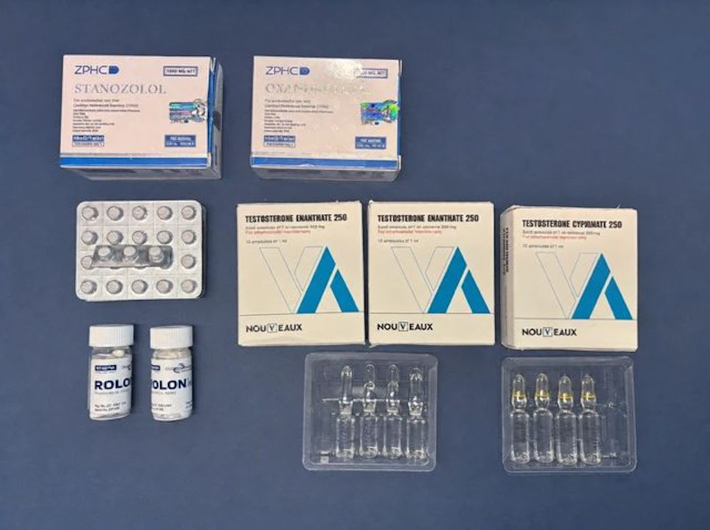 Diverse confezioni e blister di steroidi anabolizzanti, preparati di testosterone e compresse sono disposti su una superficie blu. I prodotti includono Stanozolol, Oxandrolone, Testosterone Enantato e altri medicinali. Si tratta di sostanze soggette a prescrizione e controllo.