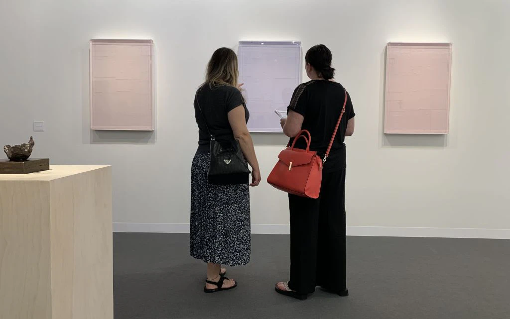 Due donne stanno in una galleria moderna davanti a tre quadri astratti di colore chiaro appesi alla parete. Una delle donne indica il quadro centrale mentre l altra porta una borsa rossa e tiene in mano un tablet. A sinistra dell immagine si vede un piedistallo chiaro con una piccola scultura.  