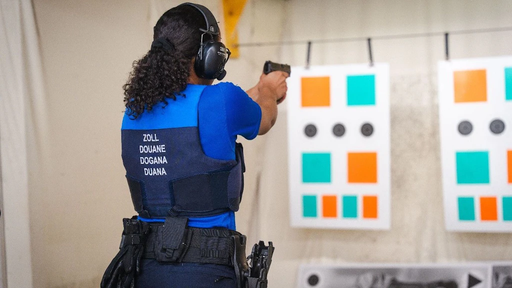 Un’agente doganale si esercita in un poligono di tiro con una pistola, mirando a bersagli colorati. Indossa un giubbotto antiproiettile con la scritta "Dogana" in varie lingue e delle cuffie antirumore. Sullo sfondo si vedono bersagli con riquadri arancioni e verdi.