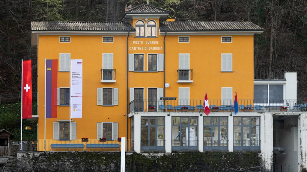 La foto mostra il museo delle dogane a Gandria, un edificio giallo che spicca con diverse bandiere, tra cui quella della Svizzera. È circondato da alberi e dispone di un balcone con sedie.