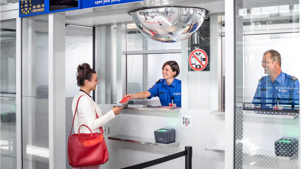 Una giovane donna con i capelli scuri appuntati, grandi orecchini d'oro, un blazer chiaro e una grande borsa rossa si trova davanti al bancone del controllo passaporti dell'aeroporto. Riceve il suo passaporto svizzero rosso da un giovane impiegato dell'UDSC. L'impiegato indossa una camicia blu corta con il logo UDSC e sorride alla donna in modo amichevole. Un dipendente UDSC con gli occhiali siede al bancone accanto e osserva la scena dalla porta accanto.