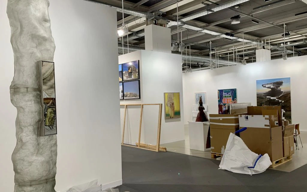 In un padiglione espositivo, pareti bianche vengono allestite con quadri e sculture mentre scatole e materiale da imballaggio sono ancora sul pavimento. La scena mostra l allestimento non ancora terminato di una fiera o mostra d arte.  