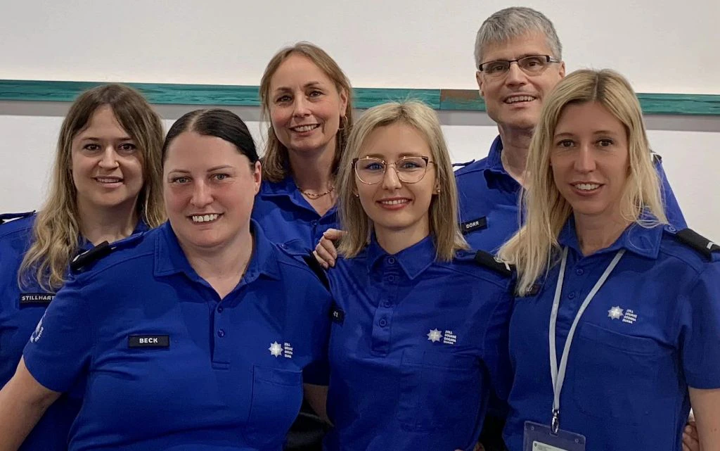 Sei collaboratori dell amministrazione delle dogane, in uniforme blu, posano fianco a fianco sorridendo verso la camera. Il gruppo rappresenta una squadra mista e affiatata.  