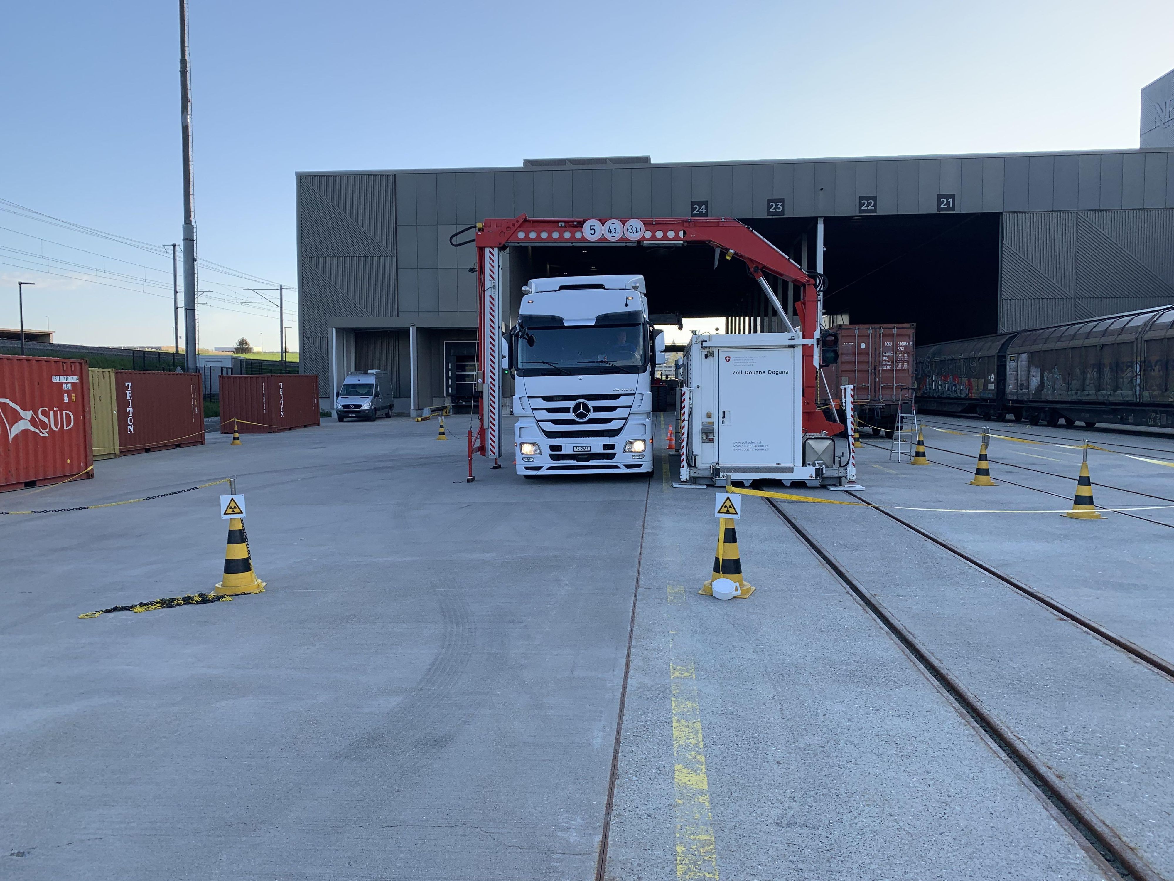 Un container bianco con un braccio di gru rosso si trova su un'area asfaltata. Di fronte al container si trova la base del braccio della gru, che funge da cancello per i camion. Un camion bianco, attualmente sottoposto a scansione, sta attraversando questo cancello. Intorno allo scanner sono posizionati dei coni neri e gialli per il traffico, mentre sullo sfondo si intravede un edificio.