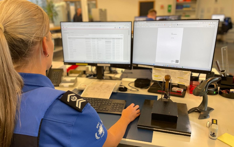 Un’agente doganale è seduta a una scrivania con due schermi ed elabora documenti elettronici. Gli schermi mostrano una panoramica dei dati e un modulo con codice QR.