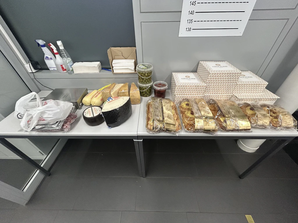 Su un tavolo sono visibili diversi alimenti come formaggi, salumi, olive, prodotti da forno e dolci confezionati. I prodotti sono ordinatamente disposti, probabilmente durante un controllo doganale. Sullo sfondo si vedono detergenti e tovaglioli di carta.  