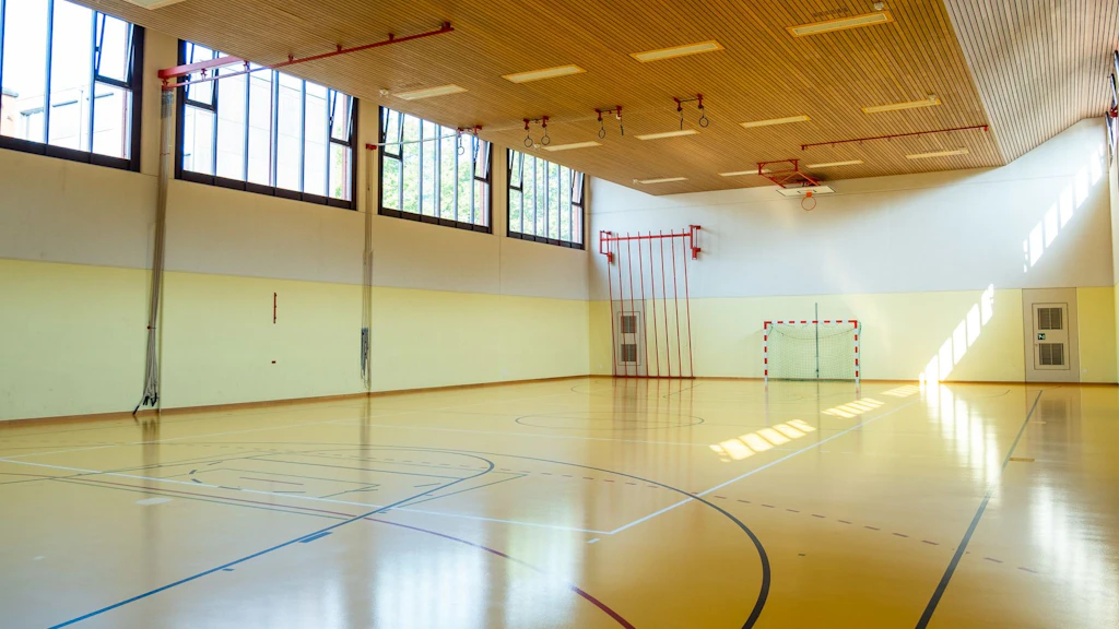 Una grande palestra luminosa con pavimento in legno e alte finestre. Sullo sfondo si vede una porta da pallamano e attrezzi sportivi sono appesi alle pareti. La palestra è vuota e ben illuminata.